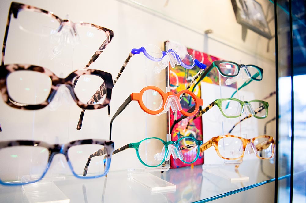 The image shows a collection of eyeglasses on display with various frame colors and styles, arranged neatly behind a glass case.