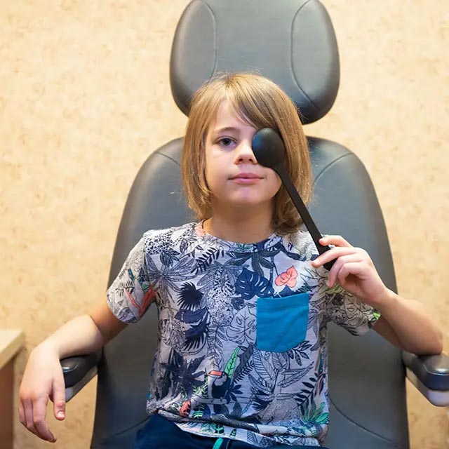 A young boy sits in an eye chair with a black spoon held up to his face, appearing to be looking through it.