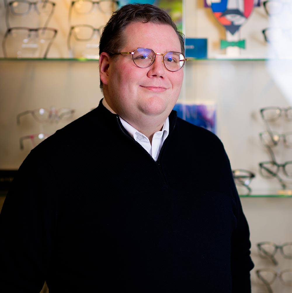 The image shows a man standing in front of a display case with glasses, wearing a dark sweater and glasses, smiling slightly at the camera.