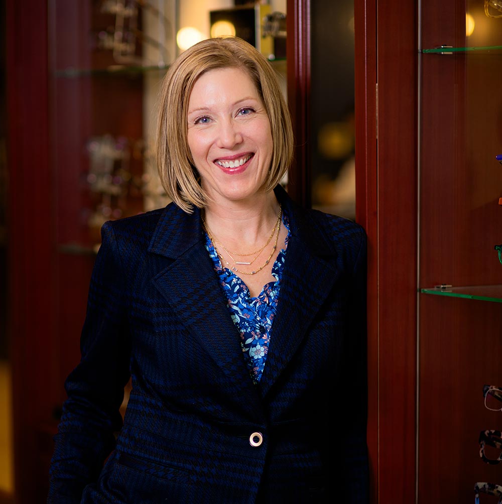 A woman with glasses and a smile stands in front of an optical store display case.