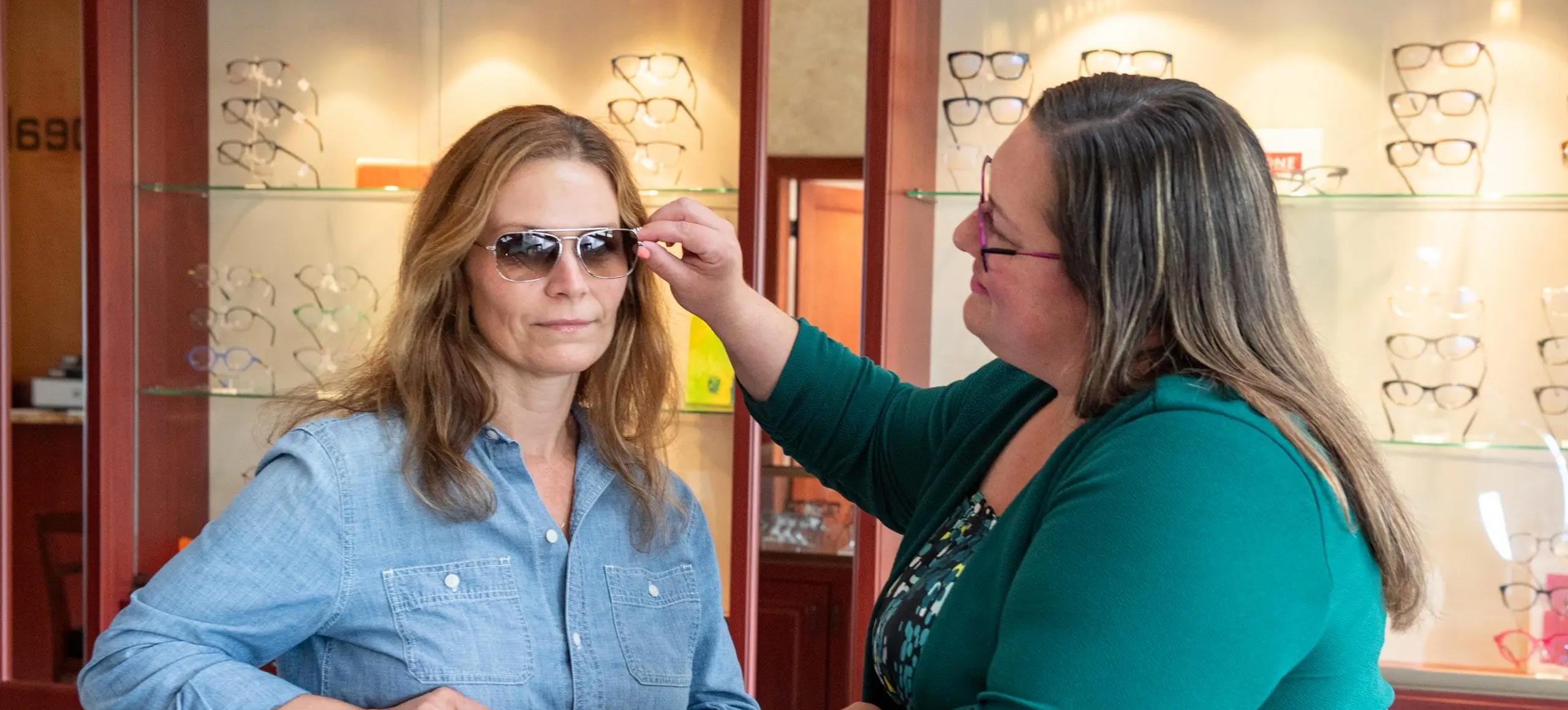 A woman is adjusting sunglasses on another woman's face at an optician shop.
