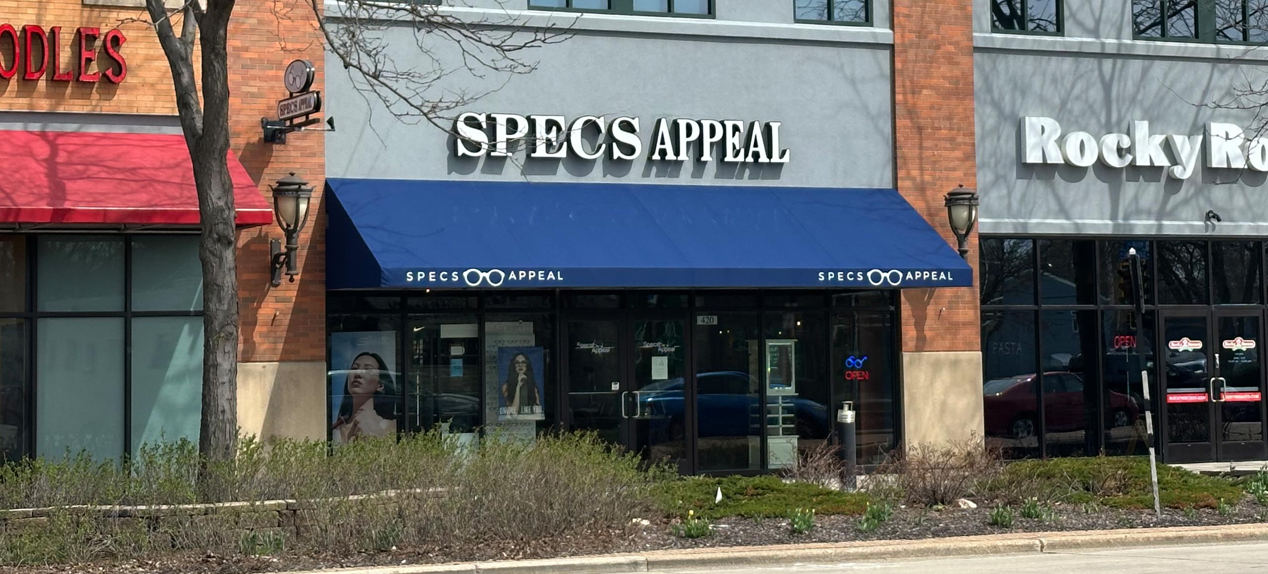 The image shows a row of storefronts with various business signs, including 'Specs Appeal' and 'Rocky Mountain Chocolate Factory,' under a clear sky on what appears to be a sunny day.