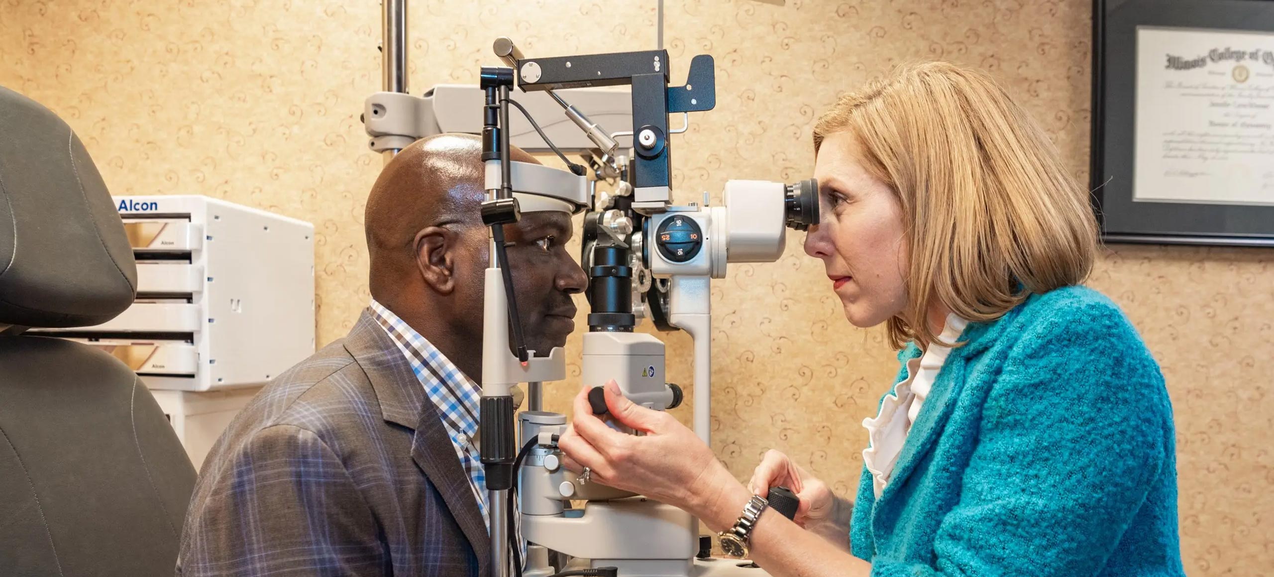 A woman in a blue coat using an eye machine on a man wearing glasses, who is seated and facing her.