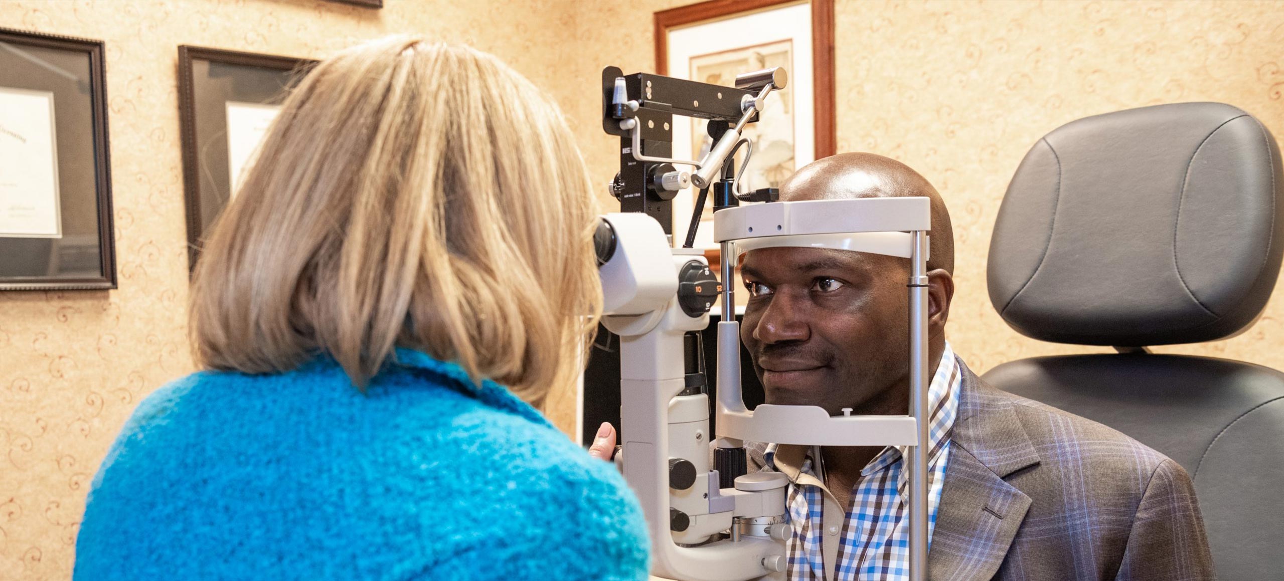 The image depicts a man sitting in an eye examination chair with his eyes dilated, being examined by a woman who appears to be an optometrist or ophthalmologist.