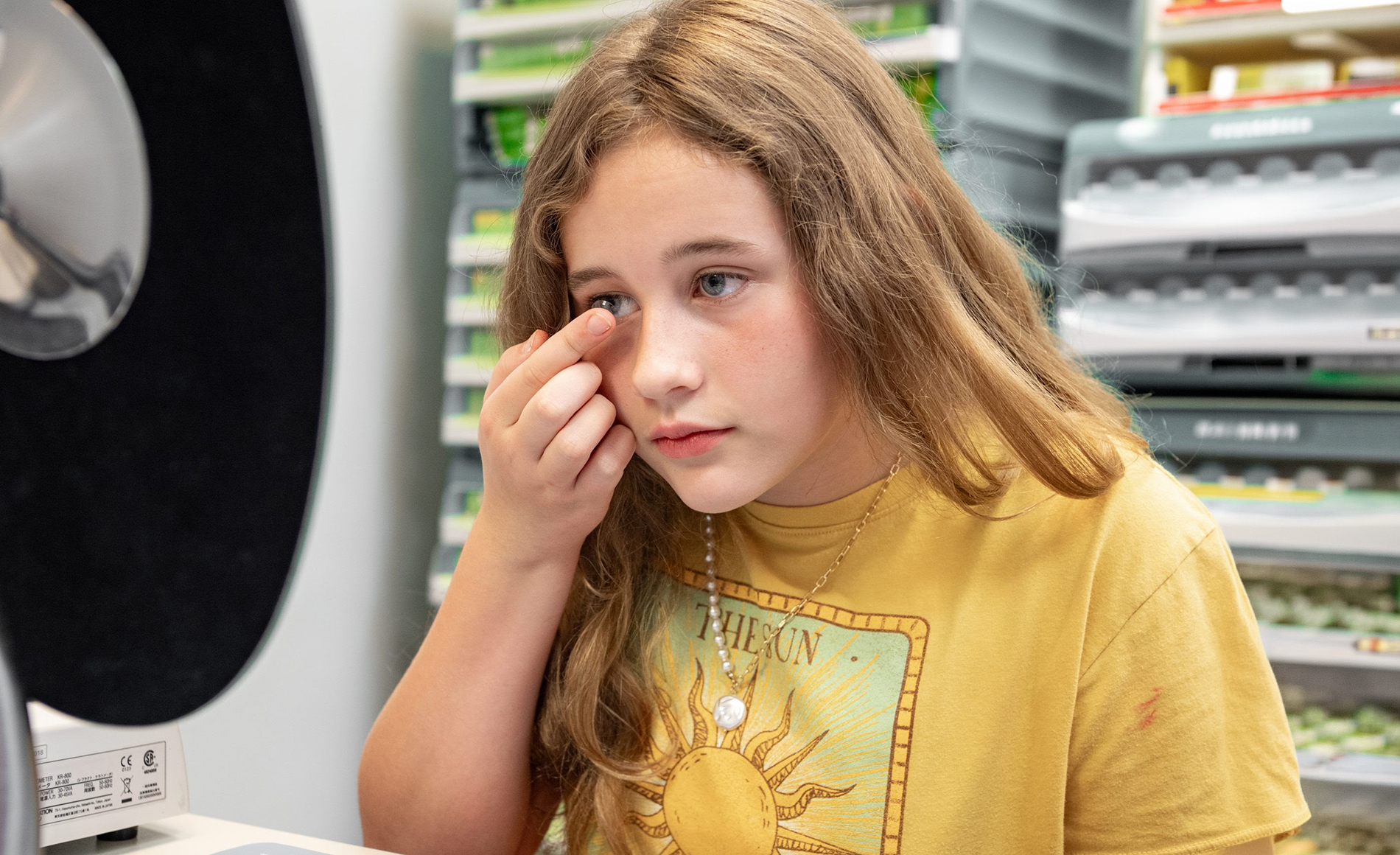 A young girl with long hair, wearing a yellow shirt, sitting at a counter in a store, holding her hand to her face while looking down, with shelves of products behind her.