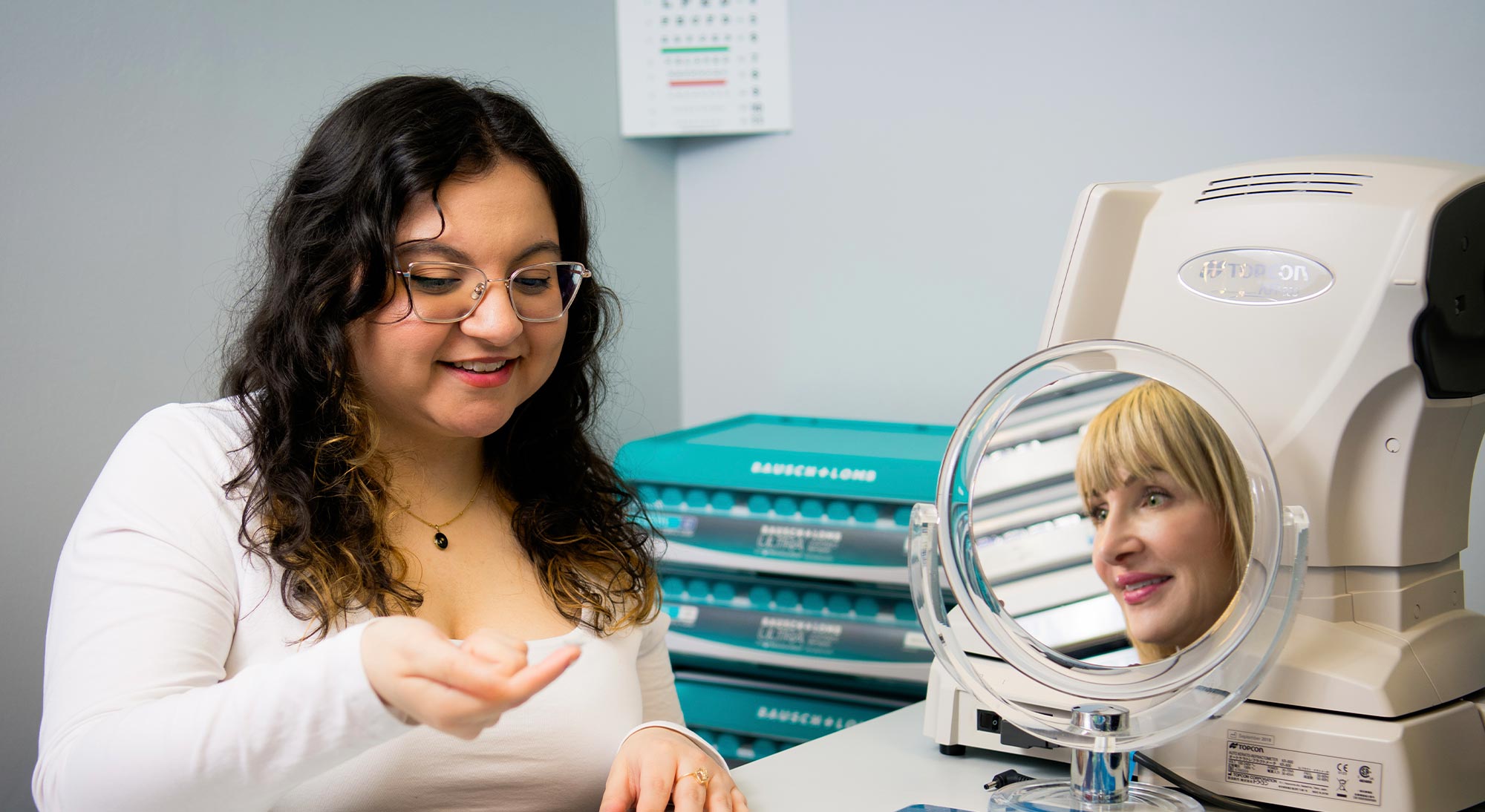 A woman wearing glasses is seated at an optical device, assisting another woman who is looking into a magnified mirror, both are in a room with medical equipment.