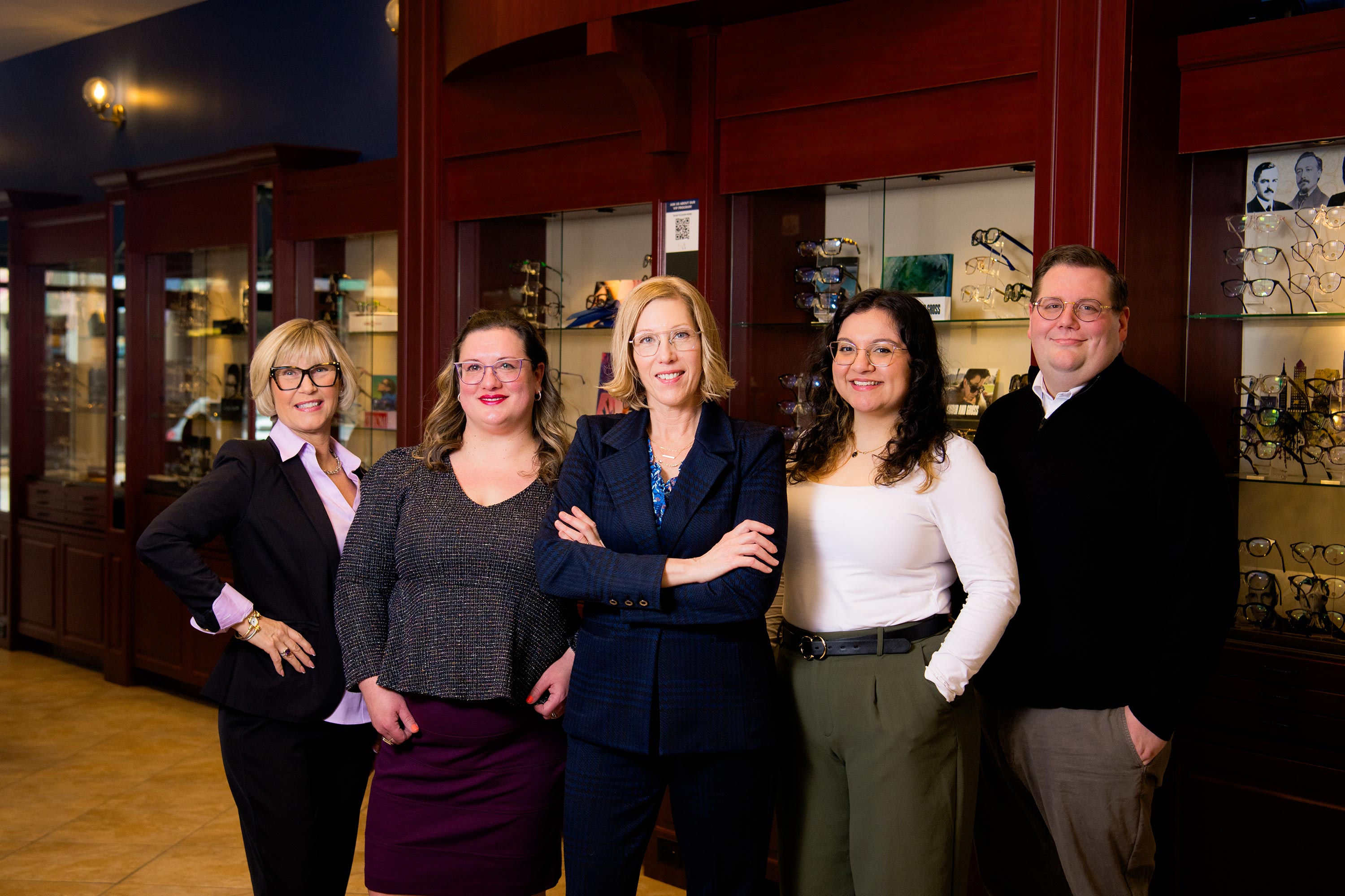 The image shows five individuals posing together inside a store with display cases containing various items, likely optical frames, in the background.