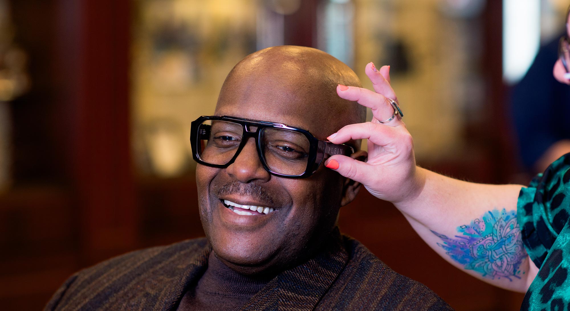 A man with glasses and a bald head being groomed by another person, likely in a barbershop setting.