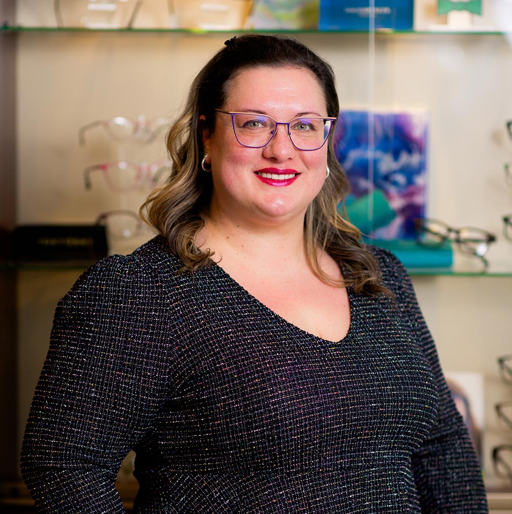 The image shows a woman standing in front of a display of eyeglasses, wearing glasses herself, with her hair pulled back and a smile on her face.