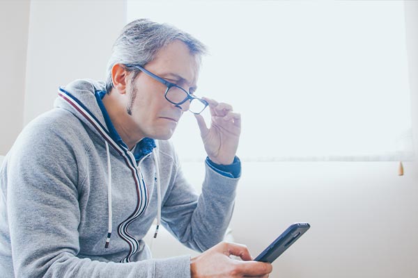 The image is a split-screen photograph showing two different scenes  On the left, a man is seated at a table with his head bowed down, presumably reading or looking at something on the table  he is wearing glasses and has a beard. On the right, another man is standing in front of a window, holding a cell phone to his ear as if engaged in a conversation. Both men are dressed casually and appear to be middle-aged or older.