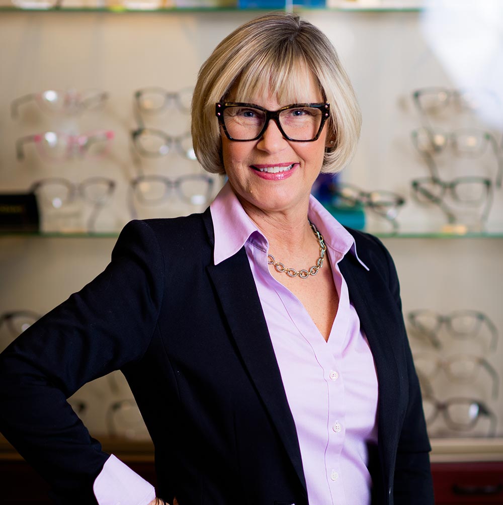A woman wearing glasses and a purple shirt stands behind a display case filled with eyeglasses.
