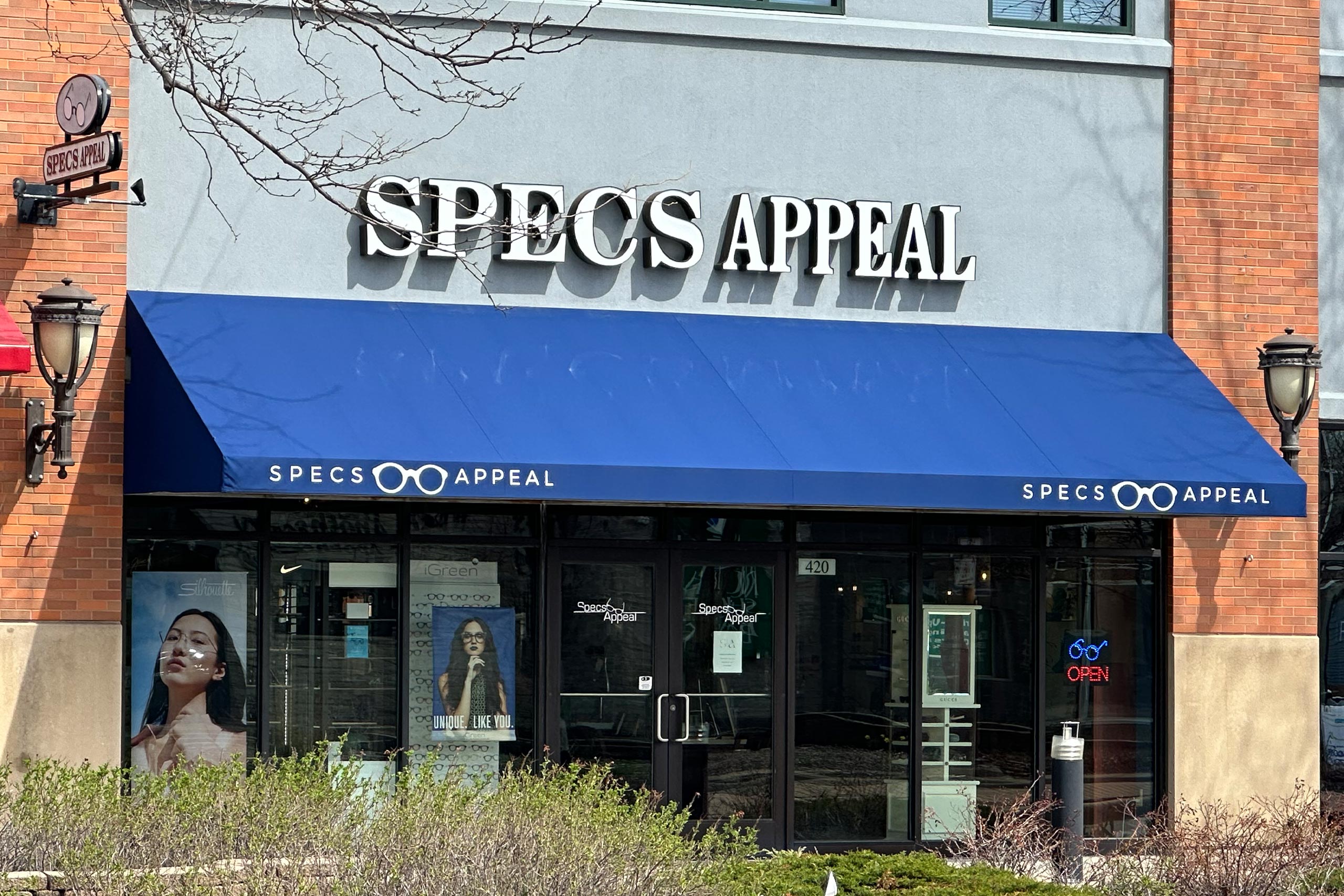 The image shows a storefront with a blue awning that reads 'SPECS APPEAL' and has a sign above it indicating it's an optical shop, situated on a street corner with a tree in the background.