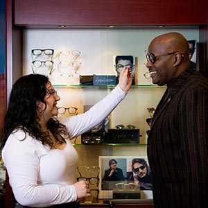 The image shows a man wearing sunglasses standing next to a woman who is applying makeup to his face. They are both inside a store with eyeglass displays in the background.