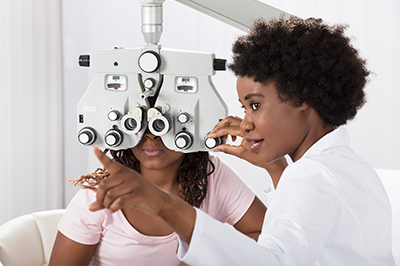 The image shows a woman sitting in front of an optometrist s eye exam machine, with her eyes dilated for a test, while the optometrist stands behind her, likely conducting the examination.