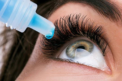 The image shows a close-up of an eye being treated with a dropper filled with a blue liquid, positioned over the eye, with the person s eyelashes visible and the focus on the eye treatment process.